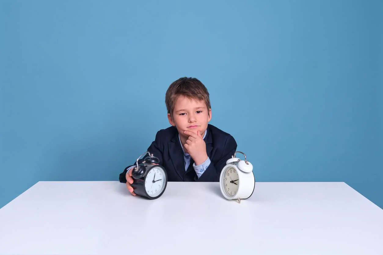 The young boy showcases his fluency in telling time as he confidently sits at a table, engrossed in studying the two clocks before him.