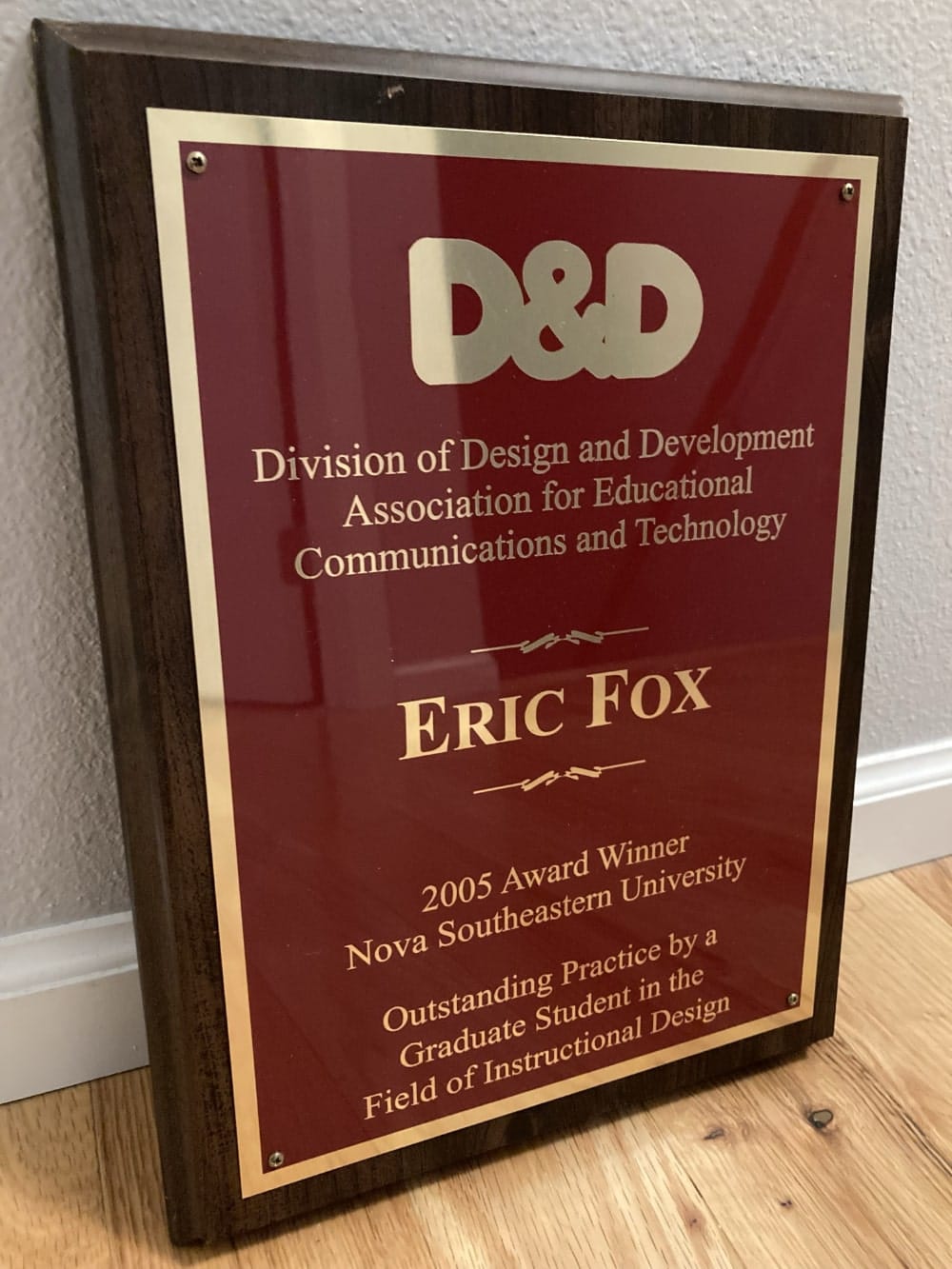 A wooden plaque with a red and gold plate awarded to Eric Fox in 2005 by the Division of Design and Development for outstanding graduate practice in instructional design, highlighting his innovative tutorial on Relational Frame Theory.